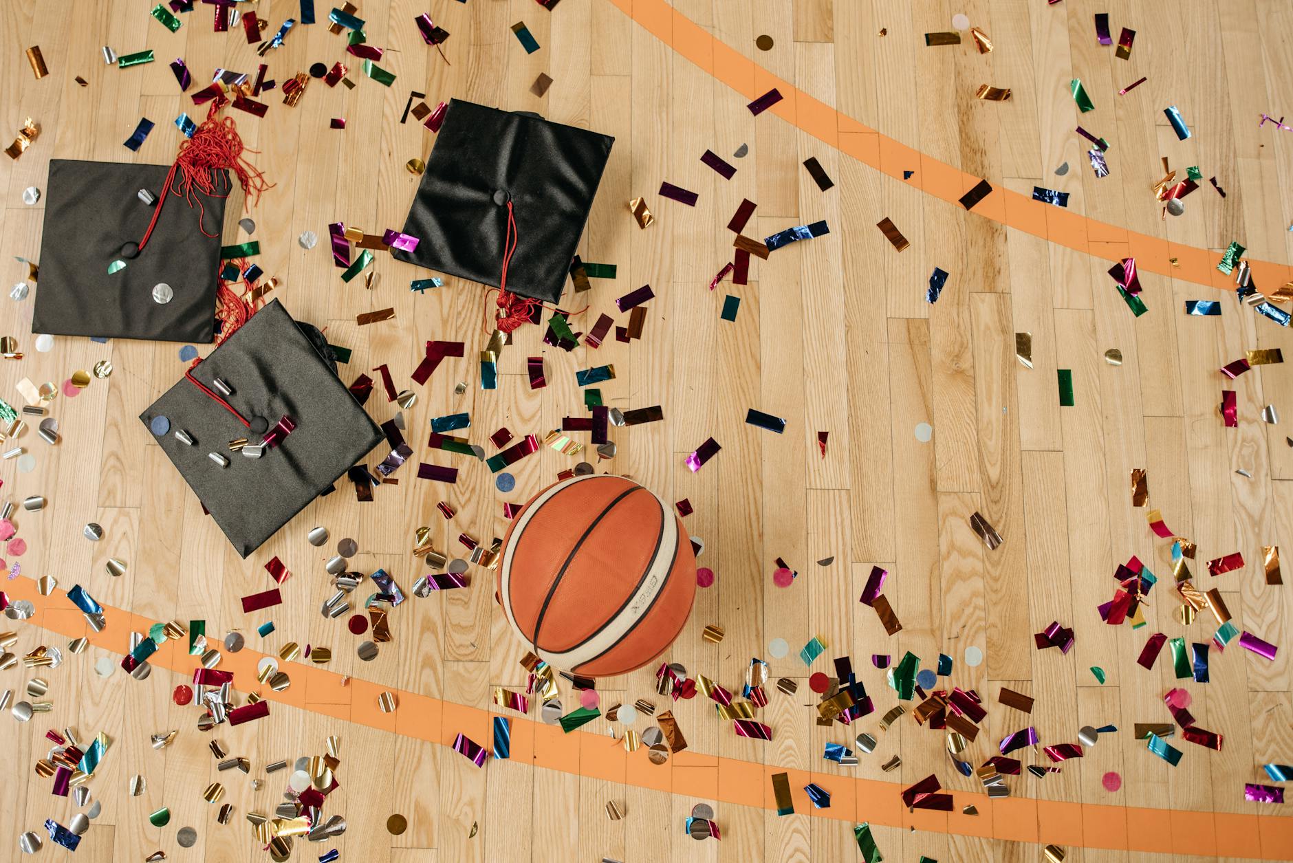 Top view of graduation caps and basketball on confetti-sprinkled gym floor.