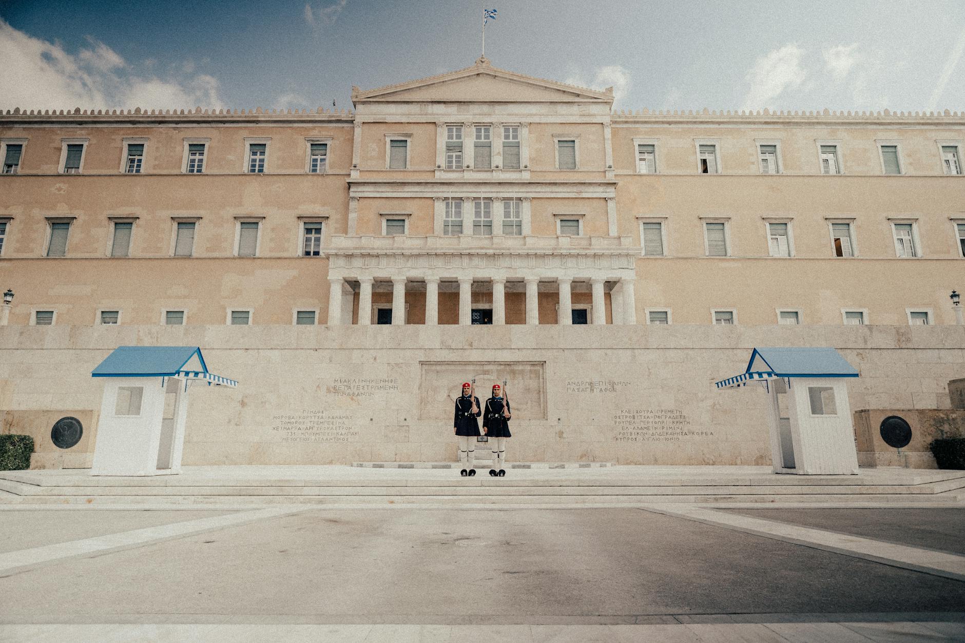 Two ceremonial guards at the Hellenic Parliament in Athens, Greece, under a blue sky.