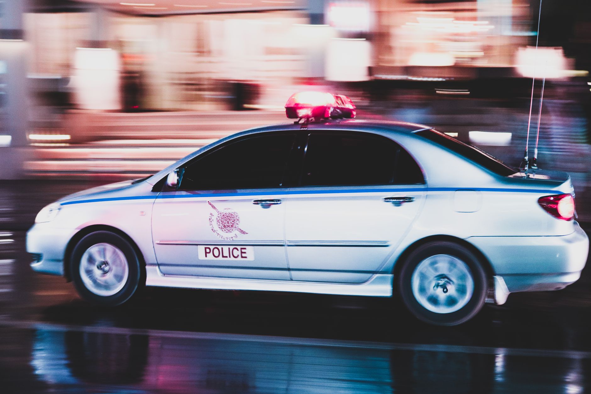 A motion-blurred police car speeds through a busy street in Thailand at night.