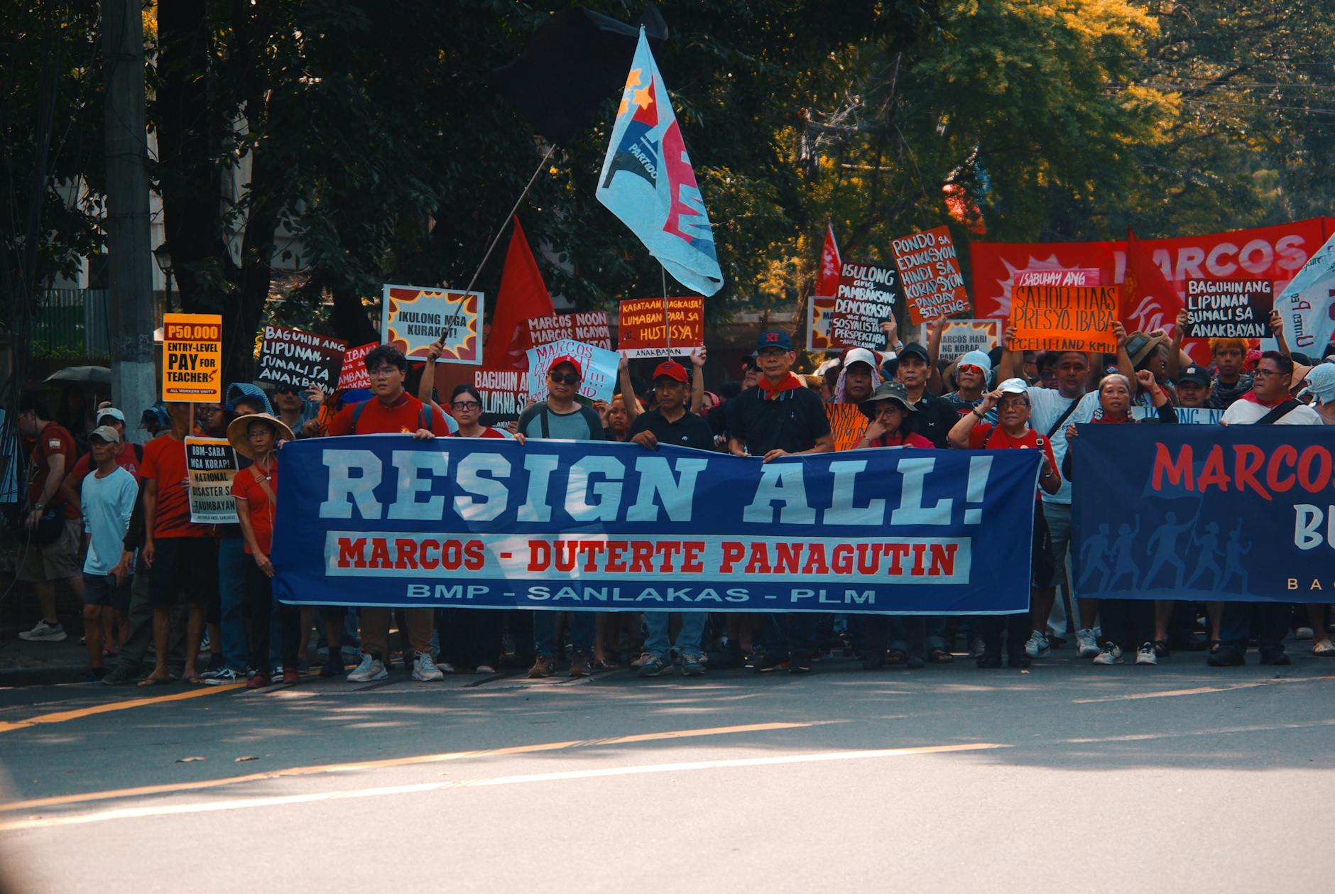 Crowd gathers in street protest holding signs demanding resignation with political messages.