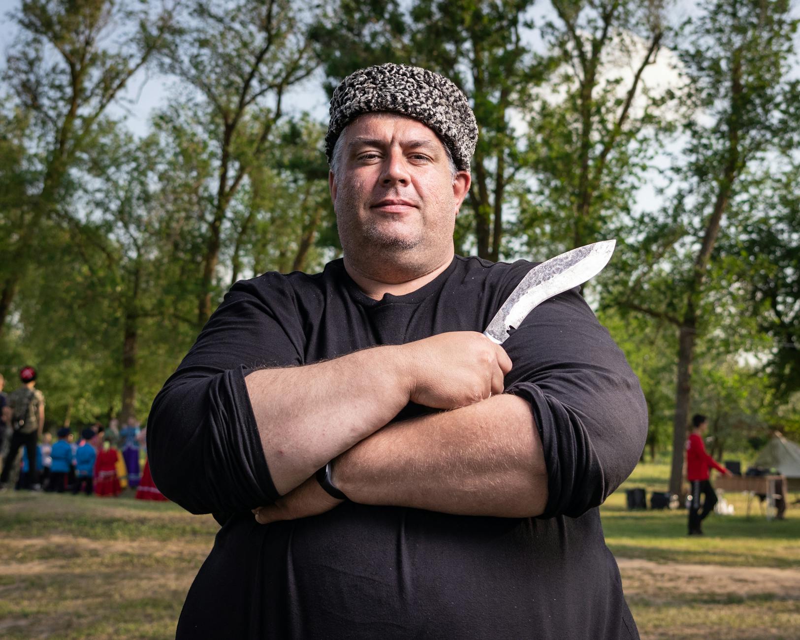 A man stands confidently outdoors holding a traditional knife, surrounded by trees.