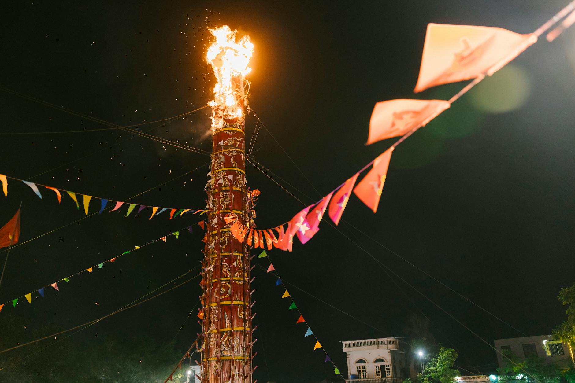 Tall flaming tower during night festival with colorful flags and people climbing.