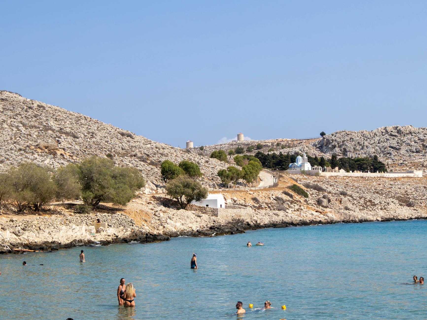 People enjoying a swim at a picturesque beach in Greece with a rocky hillside and clear blue water.
